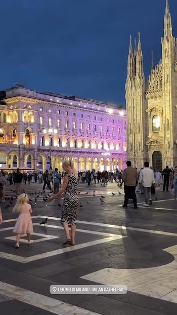 Una mujer rubia con vestido estampado y una niña con vestido rosa caminan entre palomas frente a la catedral del Duomo de Milán