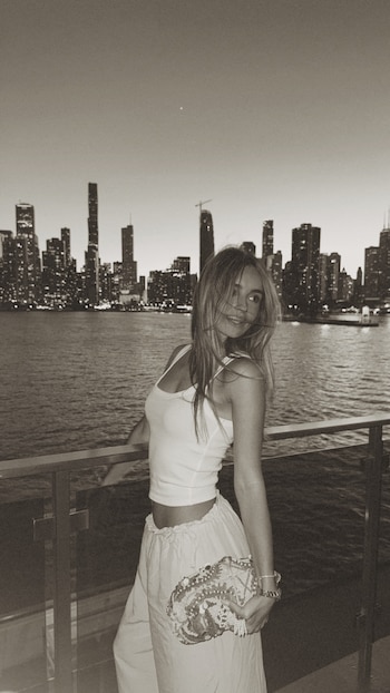 Joven mujer rubia sonríe con cartera decorada, parada junto a baranda con ciudad iluminada y agua al fondo al atardecer en blanco y negro