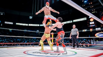Tres luchadores enmascarados realizan una llave acrobática en el centro de un ring de lucha libre junto a un árbitro con uniforme de rayas.