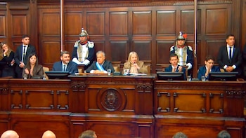 Una sala con paneles de madera oscura donde se ven varias personas sentadas y de pie en una mesa principal, flanqueados por dos guardias con uniformes ceremoniales