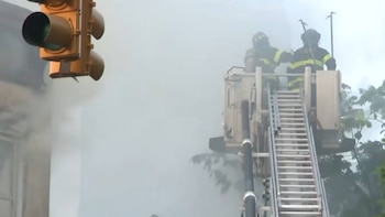 Dos bomberos en equipo de protección, de pie en la plataforma de una escalera de camión entre denso humo. Un semáforo amarillo cuelga a la izquierda