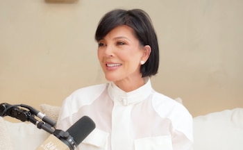 Primer plano de Kris Jenner con pelo corto oscuro, sonriendo, vestida con una blusa blanca de cuello alto. Un micrófono negro está en primer plano a la izquierda
