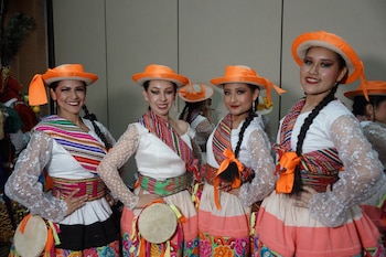 Cuatro mujeres con sombreros anaranjados, blusas blancas de encaje, faldas con bordados florales y sashes de colores, dos sostienen tambores pequeños