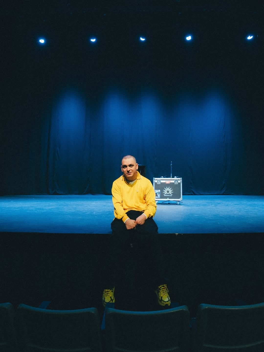 Lucho Miranda, el comediante chileno, se sienta en el escenario con luces azules de fondo, reflejando su inspiradora trayectoria y su carisma único.