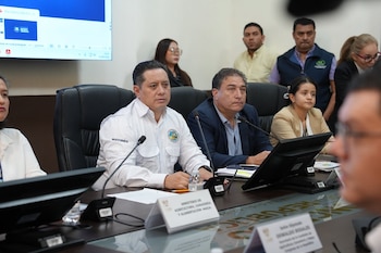 Varias personas sentadas ante una mesa con micrófonos, monitores y placas de identificación de instituciones públicas
