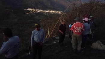 Varias personas, algunas con gorras de policía, de pie en un terreno oscuro con vegetación seca. Al fondo, se observan las luces de un poblado