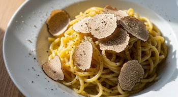 Plato de cerámica blanca con pasta tajarin amarilla, cubierta con láminas finas de trufa blanca y pimienta negra, sobre una mesa de madera rústica, iluminado con luz natural.