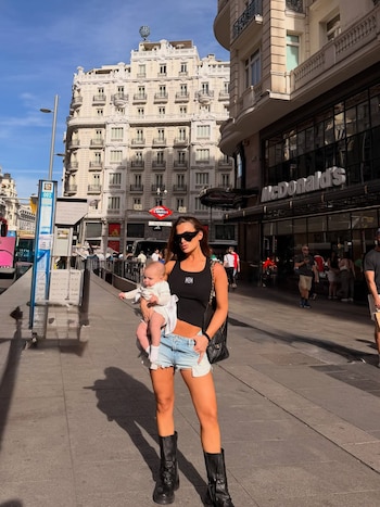 Cami Homs, con gafas de sol, top negro y shorts de mezclilla, sostiene a su bebé Aitana vestida de blanco en una acera de Madrid. Edificios y un McDonald's al fondo