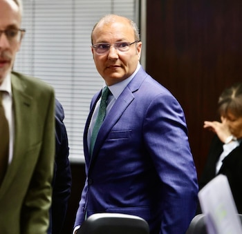 Un hombre con traje azul, gafas y corbata verde observa hacia un lado en un espacio de oficina
