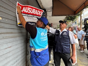 Dos inspectores municipales pegan un cartel rojo y blanco con la palabra clausurado sobre la persiana metálica cerrada de un local