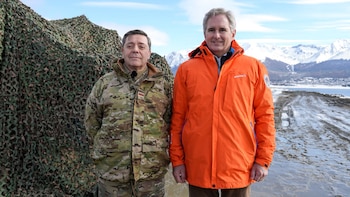 Dos hombres de pie al aire libre, uno con uniforme camuflado y otro con abrigo naranja, frente a una red militar y montañas nevadas