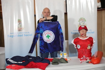 Un hombre sostiene una camiseta azul de salvavidas con el logo 'Salvamento IGSS' junto a un maniquí de RCP con camiseta roja, gorra y silbato. Hay equipo de rescate en la mesa