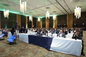Amplio salón de convenciones con cientos de asistentes sentados en mesas blancas y azules, cámaras de video, pantallas de monitoreo y luces colgantes