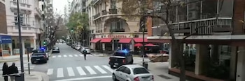 Vista de una intersección urbana con dos patrullas policiales con balizas azules y cinta de seguridad. Personas en la acera y un paso de cebra visible