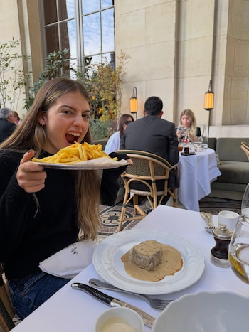 Una joven sostiene un plato con papas fritas cerca de su boca frente a una mesa con un plato de carne con salsa en un restaurante