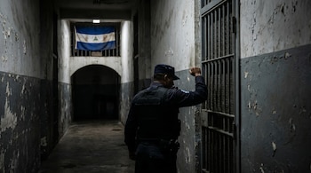 Oficial de policía de espaldas golpea una puerta de celda con barrotes en un pasillo oscuro. Paredes con pintura descascarada y bandera de Nicaragua al fondo.