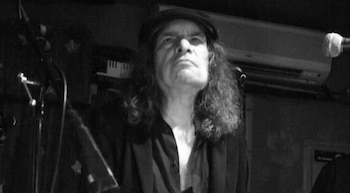 Primer plano en blanco y negro de Chet Mazur con gorra y cabello largo, mirando hacia arriba, con un micrófono y teclado al fondo