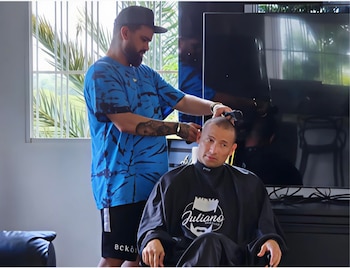 Un barbero con gorra y camisa azul corta el cabello de Cheremo, quien viste una capa negra con el logo 'Juliano', en un interior con ventana y televisor