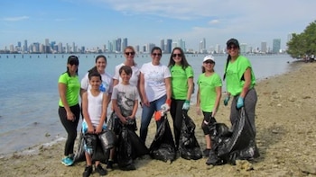 Un grupo de adultos y niños posa en una playa arenosa con bolsas de basura negras. El agua y un horizonte urbano con rascacielos son visibles al fondo
