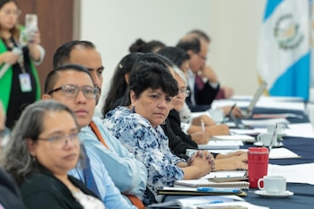 Varias personas sentadas a una mesa larga. Sobre la mesa hay cuadernos, bolígrafos, laptops y tazas. La bandera de Guatemala se ve al fondo
