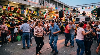Personas reunidas en una calle decorada frente al Mercado de Jamaica con arreglos florales y un escenario.