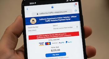 Una mano sostiene un smartphone mostrando un sitio web fraudulento de pago de multas de tráfico de California con la URL 'california-traffic-violations.com' y un total de $235.00.