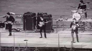 Fotografía en blanco y negro de cuatro músicos en un escenario con guitarras, amplificadores y una batería con el nombre The Beatles
