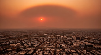 Vista aérea de un paisaje urbano con edificios y carreteras bajo un cielo naranja rojizo. El sol es un círculo brillante y el aire crea una distorsión ondulada.