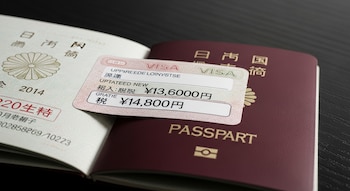 Un pasaporte japonés abierto y otro cerrado, con una etiqueta de visado sobre ellos que muestra los valores en yenes de 13.600 y 14.800.
