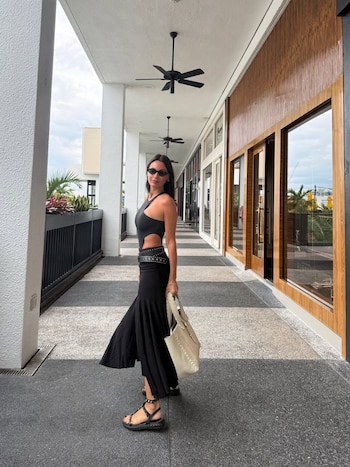 Una mujer morena con gafas de sol oscuras, top negro de un hombro y falda larga negra, camina por un pasillo exterior con tiendas, llevando un bolso de mano claro