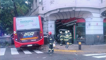 Colectivo rojo y patrullero chocados contra un edificio. Bomberos trabajan en la escena con escombros en la acera y calle. Daños visibles en la fachada