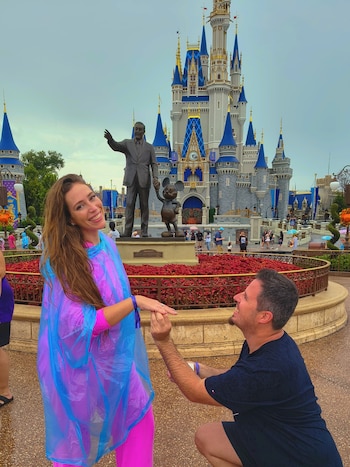 Un hombre de rodillas toma la mano de una mujer frente a la estatua de Walt Disney y el castillo de Cenicienta