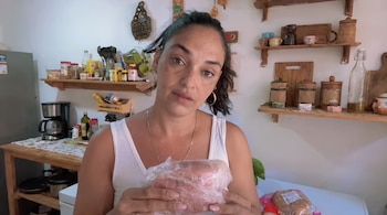 Una mujer con camiseta blanca sostiene un paquete envuelto en plástico en una cocina con repisas de madera y diversos recipientes.