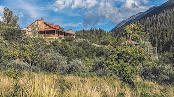 Una casa de madera con techo rojo en una colina verde rodeada de árboles frutales, pinos y vegetación densa. Al fondo, una montaña y cielo azul con nubes.