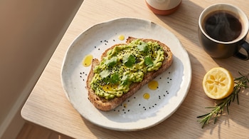 Vista aérea de una tostada de pan integral con aguacate, sésamo y cilantro en un plato blanco, junto a café humeante, medio limón y romero sobre una mesa de madera clara.