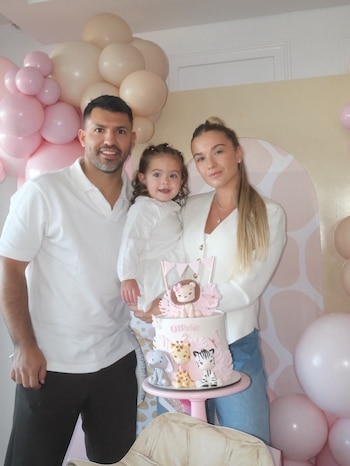 Un hombre, una mujer y una niña sonríen junto a un pastel de cumpleaños decorado con figuras de animales y globos rosados