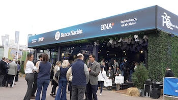 Stand de Banco Nación, logo BNA+, texto "Todo tu banco, en el celular". Follaje verde. Grupo de personas, algunas con bolsas BNA. Equipos de sonido y heno