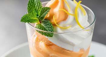 Copa de vidrio con helado bicolor, de color naranja melón y blanco, decorado con hojas de menta verde y ralladura de cáscara de limón amarilla.