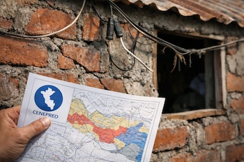 Una mano sostiene un mapa de riesgo del CENEPRED con el logo de Perú. Detrás se ve una pared de ladrillo sin revocar, cables eléctricos sueltos y una ventana.