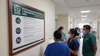 Cuatro profesionales de la salud con mascarillas, en un pasillo de hospital, observan un cartel verde y blanco del IMSS con el "Protocolo de Actuación y Prevención Ebola".