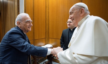 Martin Scorsese, un hombre mayor con traje azul oscuro, estrecha la mano del Papa Francisco, un hombre mayor con sotana papal blanca, ambos sonriendo. Un sacerdote con sotana negra se encuentra detrás