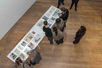 Vista aérea de varias personas de pie y agachadas observando documentos antiguos en una mesa expositora blanca, sobre un piso de madera