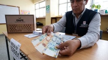 Hombre sentado de frente, con chaleco y camisa, sujeta billetes de 100, 50 y 10 soles junto a una moneda de 5 soles. En la mesa hay una caja ONPE y una identificación de mesa.
