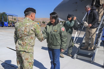 Una mujer con chaqueta verde y un hombre con uniforme militar se dan la mano en un aeródromo, con un avión y otras personas al fondo