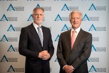 Dos hombres con traje y corbata, uno azul y otro rosa, posan frente a una pared blanca decorada con los logotipos azules de la Asociación Empresaria Argentina