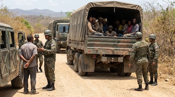 Soldados del Ejército de República Dominicana interactúan con un hombre junto a un vehículo militar, mientras un camión transporta personas en una carretera de tierra.