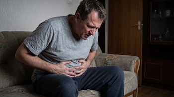 Hombre con camiseta gris y pantalón oscuro sentado en un sofá, con las manos sobre el abdomen y expresión de dolor.