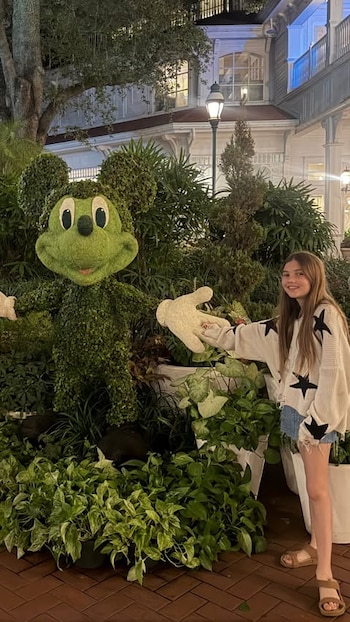 Una niña con un suéter decorado con estrellas sostiene la mano de una escultura vegetal con la forma de Mickey Mouse