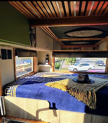 Interior de habitación con estructura de cama, colchón, manta azul, sombrero, dos ventanas con vista a una calle con vehículos y árboles, techo de madera
