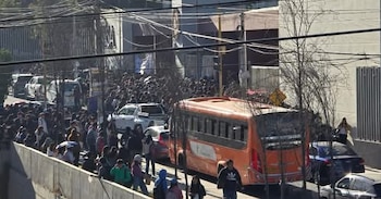 Multitud de personas en una calle con un autobús naranja y varios coches estacionados. Un edificio moderno se ve al fondo y cables eléctricos cruzan la imagen